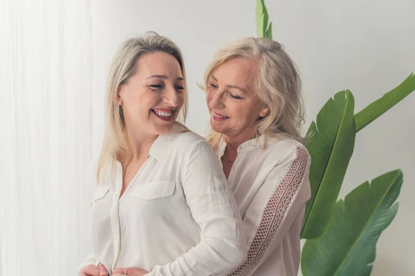 One woman with wavy blonde hair standing in the back hugging another middle-aged caucasian woman. Both laughing and having fun in an elegant apartment. High quality photo