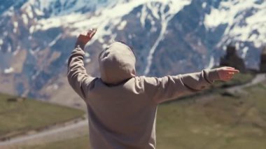 Özgürlük ve doğa kavramı. Gri kapüşonlu genç adam kollarını sallıyor ve nefes kesici dağ manzarasının üzerinde barış işareti gösteriyor. Yüksek kalite 4k görüntü