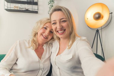 Modern bir dairede oturmuş selfie çekiyormuş gibi yapan beyaz tişörtlü, orta yaşlı, zarif iki kadının kapalı çekimleri. Yüksek kalite fotoğraf