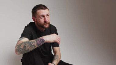 Dövmeli kolları olan yakışıklı beyaz adam sakalına dokunuyor ve yüzünü çeviriyor, gri arka plan stüdyosu orta boy fotokopi çekiyor. Yüksek kalite 4k görüntü