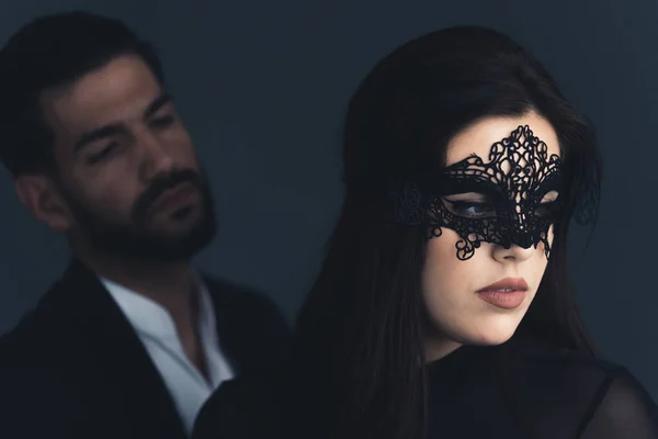 Attractive young european brunette woman in black outfit and a lacy carnival mask looking away. Elegant man standing behind her. Focus on the foreground. Studio shot. High quality photo