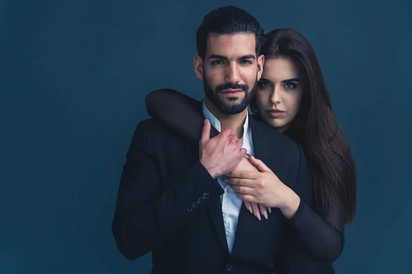 Medium studio shot over dark blue background of a sensual interracial couple wearing elegant clothes, woman hugging the man from the back. High quality photo