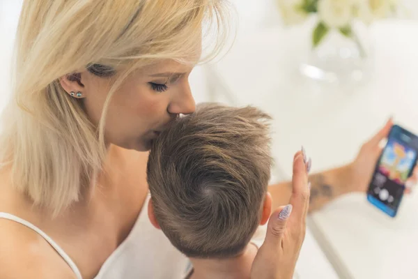 mom kissing her little son on the head and watching something in the phone with him. High quality photo