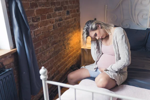 Health problems during pregnancy. Worried caucasian blonde pregnant middle-aged woman sitting on a bed and contemplating about her uncertain future. High quality photo