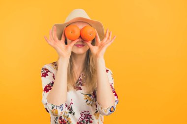Sağlıklı yeme alışkanlıkları konsepti. Kafkasyalı, uzun saçlı, şapkalı ve çiçekli bluzlu iki canlı portakalla gözlerini kapayan genç bir sarışın. Stüdyo, turuncu arka plan. Yüksek kalite fotoğraf