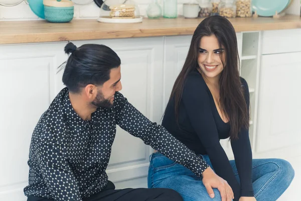 Black-haired bearded handsome man in a patterned black shirt sitting on the kitchen floor with the beautiful long-haired girl he is dating. Happy relationship. High quality photo