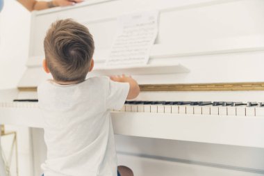 Little unrecognizable caucasian boy in a white t-shirt sitting in front of a beautiful ivory white piano. Music as a form of hobby for young kids. High quality photo