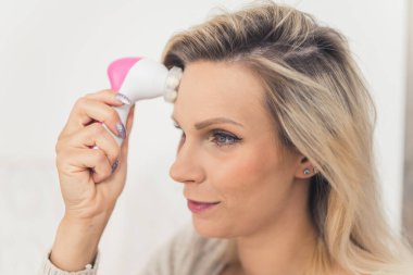 Modern cilt bakım rutini. Yüzündeki kırışıklıklardan kurtulmak için alnına otomatik masaj yapan, renkli sarı saçlı mutlu beyaz kadın. Yüksek kalite fotoğraf