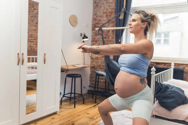 Caucasian blonde pregnant woman in her 30s wearing white biker shorts calmly doing a squat in her bedroom. Active pregnancy concept. High quality photo