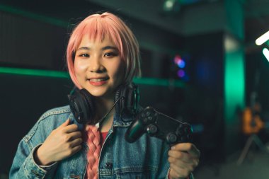 Young asian cute happy female player with pink hair and a long braid wearing headphones around her neck, holding a gamepad, smiling,. High quality photo