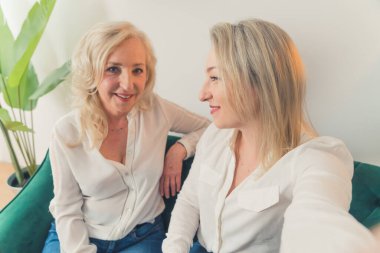 İki neşeli, beyaz saçlı, kanepede oturmuş birbirlerine bakıp gülümseyen, beyaz şık gömlekli iki kadının olduğu bir dairede çekilmiş bir fotoğraf. Yüksek kalite fotoğraf