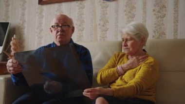 Endişeli emekli çift kanepede oturuyor ve röntgen sonuçlarına bakıyor. Orta boy kapalı mekan hastalığı konsepti. Yüksek kalite 4k görüntü