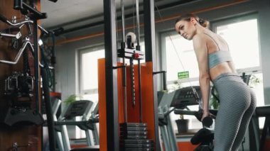 Indoor gym shot of a skinny caucasian young adult woman with slicked back hair trying out professional gym equipment for the first time. Arms exercises. High quality 4k footage