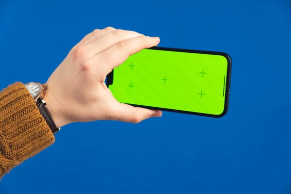 Hand holding the smartphone with green screen chroma key vertically over the blue screen background. High quality photo