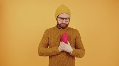 Good-looking stylish bearded man wearing mustard sweater holding an heart 3d model next to his chest - orange background. High quality 4k footage
