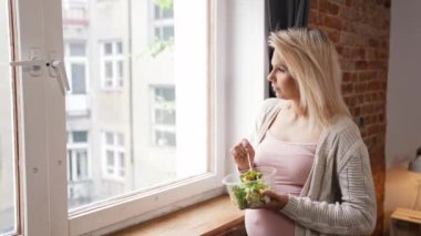 Caucasian pregnant woman with vegetable salad standing near the window, cowboy shot pregnancy and healthy food concept. High quality 4k footage