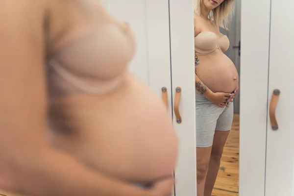 Pregnant belly in the mirror. Happy unrecognizable millennial caucasian soon-to-be mum standing in front of a mirror. High quality photo