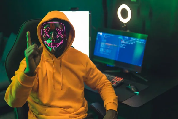 Black man wearing a dark mask with pink patterns covering his face, pointing his finger at the camera, sitting in front of a big modern computer screen. High quality photo