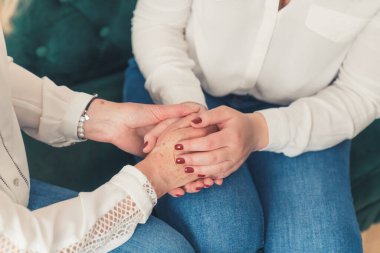 İki tanınmayan beyaz kadın kanepede el ele oturuyor. Beyaz gömlek ve kot pantolon. Kırmızı tırnaklar. Anne ve kızı. Yüksek kalite fotoğraf