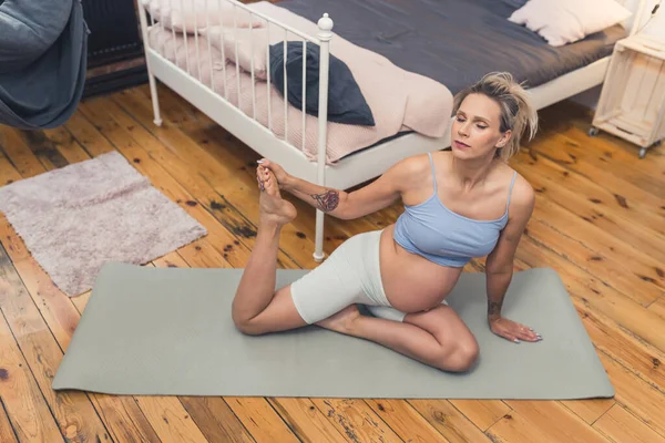 Breathing and stretching exercises for pregnant women. Young soon-to-be-mum sitting on a yoga mat with and holding one of her legs up while breathing calmly. Active pregnancy concept. High quality