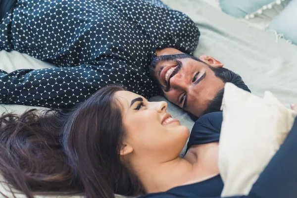 Closeup portrait photo of two young adults - handsome bearded dark-haired Cuban man in a patterned black shirt and a beautiful long-haired woman with black eyeliner - lying down together on bed