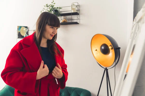 Young white brunette woman with bangs wearing a red coat looking at herself in a mirror. Indoor shot. High quality photo