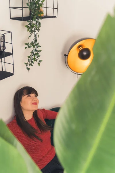 Caucasian brunette woman with bangs smiling sitting on sofa seen through leaves of house plant. Hanging plant. Focus on background. Indoor shot. High quality photo