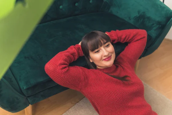 White brunette woman with bangs wearing a red sweater smiling lying on carpet resting head on arms on green sofa. Indoor shot. High quality photo