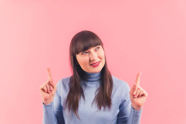 Caucasian woman with dark hair and bangs wearing blue turtleneck pointing up with both hands and looking up. Pink background studio shot. High quality photo