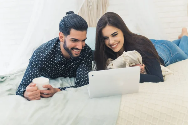 Cute couple watching film on laptop on bed with white sheets while man is holding a cup of coffee.