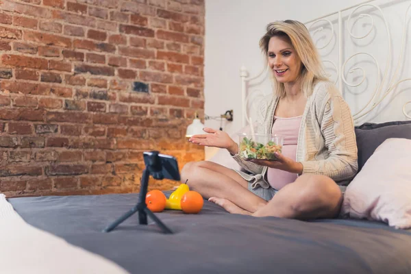Millennial caucasian influencer blonde woman sharing her pregnancy advice with her followers during livestream via smartphone. Salad. Healthy eating. High quality photo
