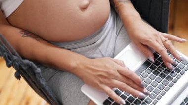 Caucasian pregnant person with long sparkly nails working on a laptop while sitting in an indoor hammock. High quality 4k footage