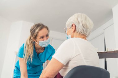 Mavi bir üniforma ve koruyucu bir maske takan genç ve güzel bir bayan doktor, bir muayene odasında yaşlı hastasıyla konuşuyor ve gülümsüyor. Yüksek kaliteli fotoğraf.