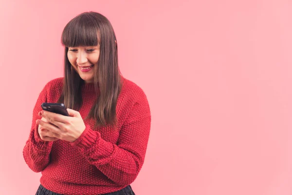 Caucasian woman with dark hair and bangs smiling wearing red sweater using smartphone. Pink background studio shot. High quality photo