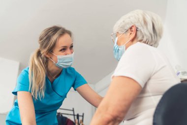 A young female medical professional, wearing a blue uniform and a face mask, having a pleasant conversation with her senior woman patient in an examination room. High quality photo