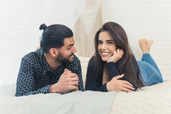 Young white couple relaxing in bedroom together. Boyfriend looking at his girlfriend whos looking at the camera with a wide smile. Indoor shot. High quality photo