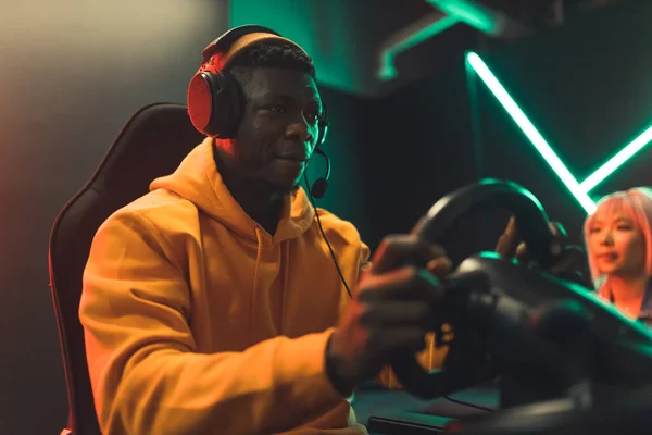 Black teenager sitting in gaming chair wearing headphones playing racing game using steering wheel. Dark red and green lighting. High quality photo
