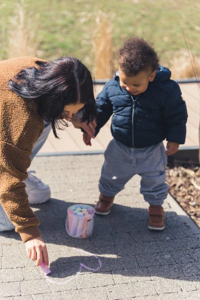 Beautiful caucasian Mother drawing a heart on the pavement using colourful crayons and her african child, both wearing winter clothing - park background. High quality photo