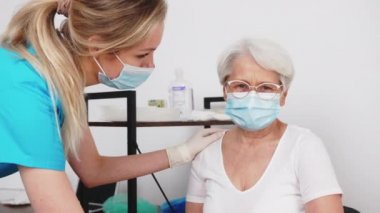 Senior lady with grey hair and her glasses on smiling sincerely at the doctor while wearing a face mask during a medical checkup. High quality 4k footage