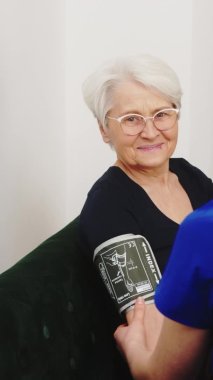 Koyu mavi üniformalı ve maske takan beyaz bir hemşire, yaşlı kadın hastasıyla kanepede oturuyor ve kan basıncını ölçüyordu. Dikey. Yüksek kalite 4k görüntü