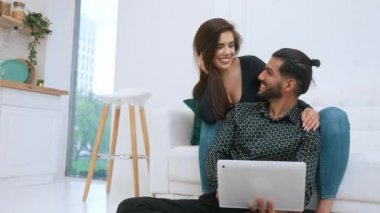 Genç çift oturma odasında dizüstü bilgisayara bakarak rahatlıyor. Beyaz kadın koltukta oturmuş gülümsüyor. Ortağına yaslanıyor ve yere oturuyor. Kapalı çekim. Yüksek kalite 4k görüntü
