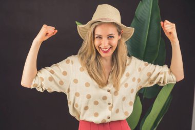 Gülümseyen, çekici, şık, şapkalı, güçlü duruşunu elleriyle gösteren bir kız. Siyah, bitki yapraklı arka plan. Yüksek kalite fotoğraf