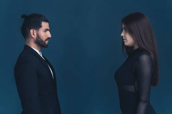 Hispanic man with hair in bun and white woman with dark long hair wearing formal clothes standing in front of each other and staring. Dark background studio shot. High quality photo