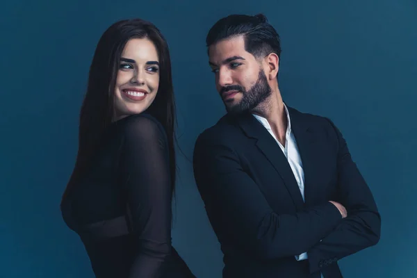 Young couple in black formal clothes standing back to back looking at each other flitring. Dark background studio shot. High quality photo