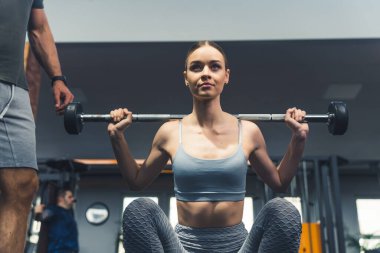 Formda kız spor salonunda ağırlık kaldırıyor ve erkek antrenör onu izliyor. Yüksek kalite fotoğraf