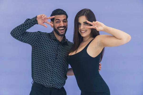 Yüzlerinde barış işareti olan, koyu renk kıyafetler giymiş gülümseyen genç bir çift. Stüdyo çekimi. Yüksek kalite fotoğraf