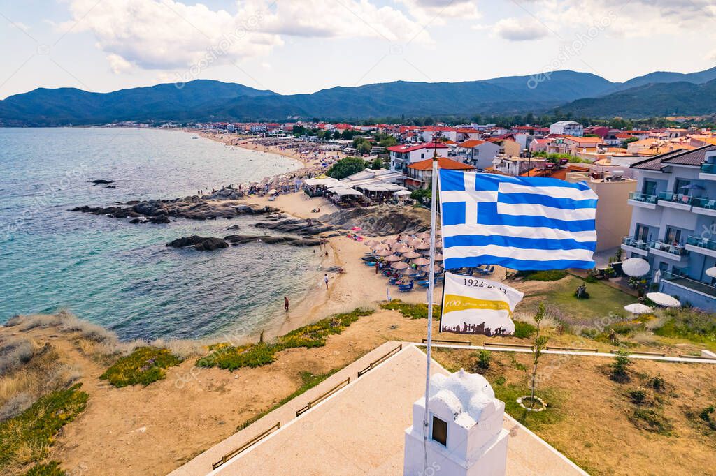 Greek city known for beachfront promenade around a rocky bay seen from aerial drone perspective. Greek flag on the foreground. High quality photo