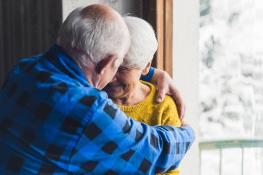 Happy loving senior husband and wife smiling and hugging near the window in the living room at home, enjoying their life together. High quality photo