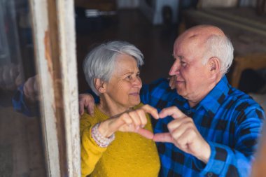 Sweet caucasian senior couple standing together, looking at each other and showing a hand heart. High quality photo