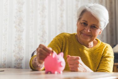 Optimistic happy smiling caucasian senior woman putting coins into a piggy bank. Savings. High quality photo